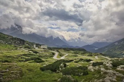 A wide shot of the St. Gotthard Pass in Switzerland, showing a winding asphalt road through green, rocky terrain. Modern white buildings stand near the road, with large mountains and dramatic clouds i
