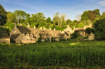 A row of historic stone cottages with pitched roofs and small windows, surrounded by green trees and a grassy field under a blue sky.