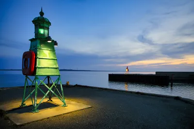 A starboard navigational light structure in the foreground with a red lifebuoy, overlooking a harbor with another port light on the sea wall in the distance at dusk.