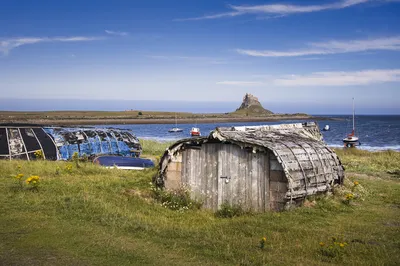 Two old, upturned boat sheds made of wood and corrugated metal on a grassy shore, with a blue sea and Lindisfarne Castle on a distant hill under a clear blue sky.