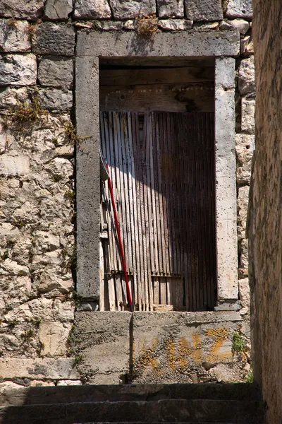 An old stone doorway with a wooden slatted interior and a red pole leaning against it, set in a narrow stone passage with steps in the foreground.