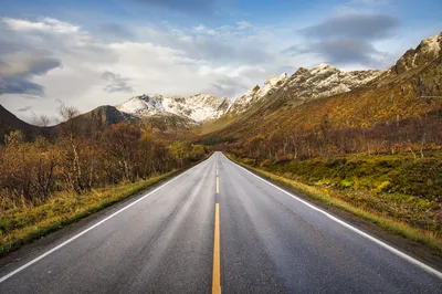 A straight asphalt road with a yellow dashed line and white solid lines on the shoulders, leading into a valley. Snow-capped mountains rise on both sides, with bare trees and autumn shrubs lining the 