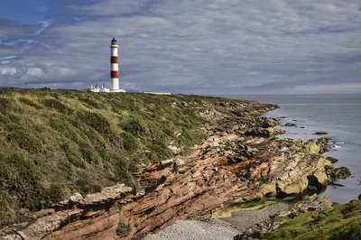 A red and white striped lighthouse on a grassy cliff overlooking a rocky coastline and the sea under a cloudy sky, with a person on the rocks.