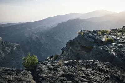 Panoramic view of a vast mountain range and deep valley at sunrise. Rugged dark rocks and sparse green bushes in the foreground, with hazy, layered mountains receding into the distance under a bright 