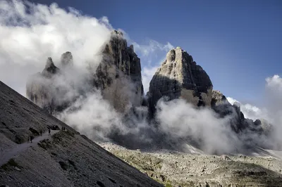 A group of hikers walk along a winding trail on a rocky mountain slope, with the jagged peaks of the Tre Cime di Lavaredo partially covered by clouds in the background.