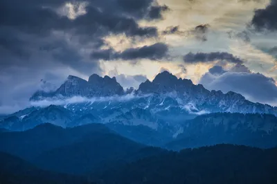 A panoramic view of rugged mountain peaks partially obscured by clouds and mist, with dark forests in the foreground and a dramatic sky at sunset.