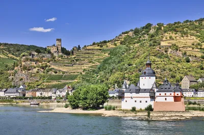 A white and red castle on a small island in a river, with a town on the bank and a ruined castle on a vineyard-covered hill under a blue sky.