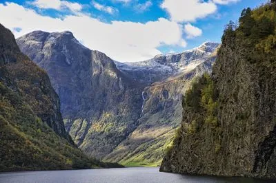 A wide shot of a Norwegian fjord with dark water in the foreground, flanked by steep, tree-covered cliffs with autumn colors. In the background, snow-capped mountains and a waterfall are visible under