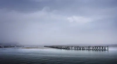A long wooden pier stretches across a misty estuary under a grey, overcast sky, with distant buildings and boats.