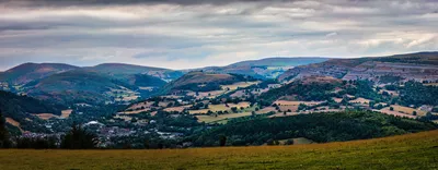 Panoramic landscape photograph of a valley with rolling green hills, a town, and a layered rock escarpment under a cloudy sky.