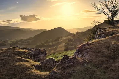 A landscape photograph of Dinas Bran Castle ruins on a hill, overlooking a valley with rolling hills and trees, illuminated by warm sunset light.