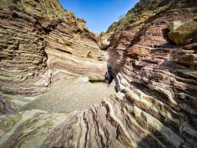 A person walks through a narrow canyon with multi-colored, horizontally layered rock walls under a clear blue sky.