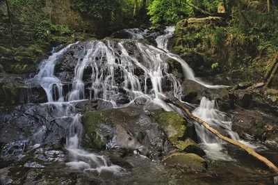 A long exposure photograph of a multi-tiered waterfall cascading over dark, moss-covered rocks in a lush green forest.