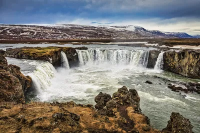 Water pours over a rocky ledge into a churning river, with snow-streaked mountains beneath a cloudy sky.