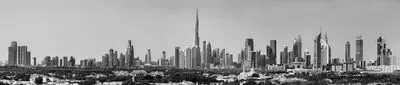 A wide panoramic black and white photograph of the Dubai skyline, featuring the very tall Burj Khalifa skyscraper surrounded by many other modern high-rise buildings under a clear sky. Trees and lower
