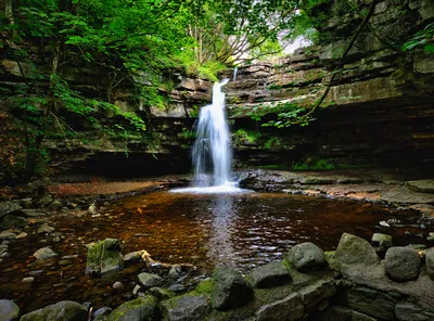 A powerful waterfall cascades into a dark, rocky pool surrounded by moss-covered cliffs and dense green trees in a shaded gorge.