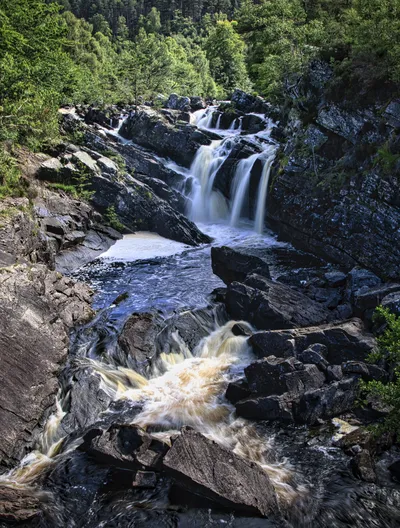 A long exposure photograph of a multi-tiered waterfall cascading over dark rocks, surrounded by green trees and foliage in a forest setting.