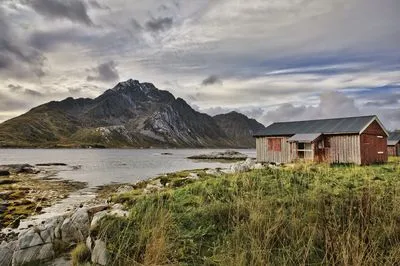 A rustic red and natural wood cabin with a dark roof sits on a rocky, grassy shore. Behind it, a calm body of water leads to large, rugged mountains with some snow on their peaks under a cloudy sky.