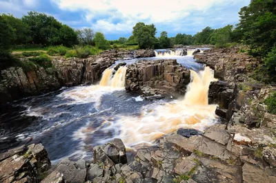 A river with several waterfalls cascading over dark, layered rock formations, surrounded by green trees under a cloudy sky.