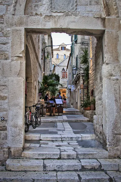 A stone archway frames a view of a narrow, stepped alleyway in Hvar, Croatia, with people dining at an outdoor restaurant and bicycles parked nearby.