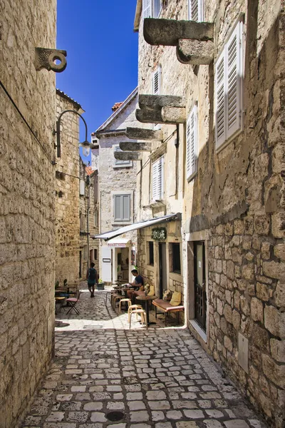 A narrow, stone-walled alleyway in Hvar, Croatia, with a cafe on the right side where people are seated at outdoor tables. Sunlight casts shadows on the cobblestone path.
