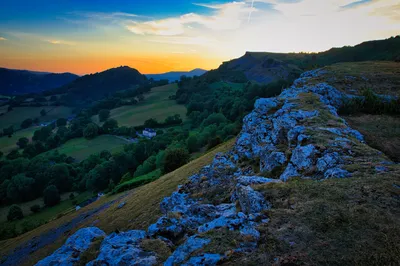 A wide landscape view of a valley at sunset, with rolling green hills, dense trees, and a distant castle ruin under an orange and blue sky. Rocky outcrops are in the foreground.