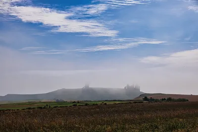 A landscape view of Dunstanburgh Castle ruins partially hidden by thick sea fog, with a green golf course and fields in the foreground under a blue sky with white clouds.