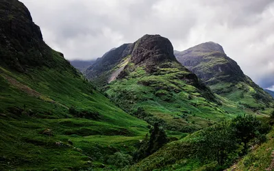 View of three large, green, rugged mountains in a valley under a cloudy sky.