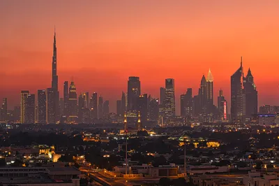 A wide shot of the Dubai skyline at sunset. The Burj Khalifa stands tall in the center-left, surrounded by numerous other skyscrapers with their lights on. The sky is a gradient of deep orange and red