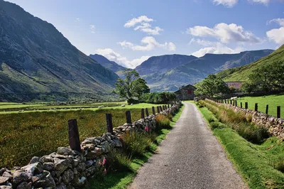 A narrow paved road winds through a green valley with steep mountains on either side, leading towards a stone building. Stone walls and fences line the road, with sheep grazing in fields.