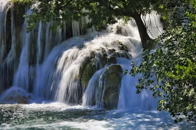 A wide waterfall cascades over multiple tiers of rocks and moss, surrounded by lush green trees and foliage, with sunlight dappling the water.