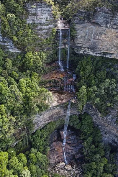 Aerial view of Katoomba Falls in the Blue Mountains, Australia. Water cascades down a multi-tiered rocky cliff face, surrounded by dense green trees and lush vegetation.