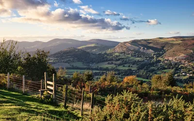 A wide landscape view of a green valley with rolling hills and scattered trees under a partly cloudy sky, with a wooden fence and stile in the foreground.