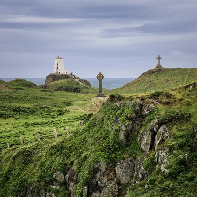 A white lighthouse on a rocky island with two stone crosses on grassy hills, overlooking the sea under a cloudy sky.