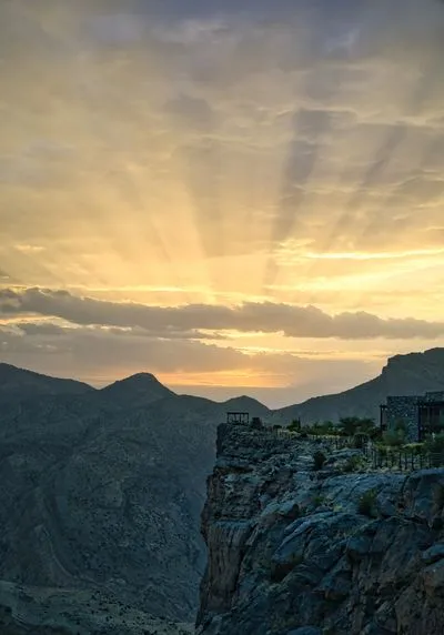 A wide shot of a sunset over mountainous terrain. Golden sunbeams radiate from behind clouds, illuminating a deep valley. A building and a small gazebo are visible on a cliff edge in the foreground.
