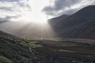 Sunlight streams through clouds over a green and brown valley flanked by dark mountains. A river winds through the valley floor, with scattered buildings and stone walls visible.