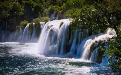 A wide waterfall with multiple tiers of white water cascading into a river, surrounded by lush green trees and foliage under bright sunlight.