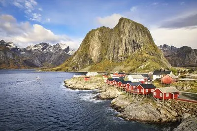 A fishing village named Hamnoy in Lofoten, Norway, featuring red houses on stilts along a rocky coastline. Snow-capped mountains rise in the background, with a boat leaving a wake in the still water.