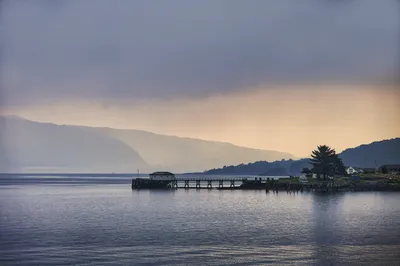 A long wooden jetty with a small building on its end stretches into a calm bay, with misty mountains in the background under a cloudy sky.