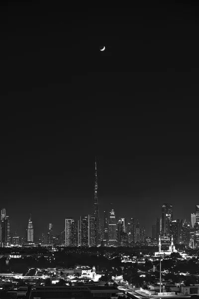 Black and white photograph of the Dubai skyline at night, with the Burj Khalifa and other skyscrapers brightly lit. A crescent moon is visible in the dark sky above the city lights.