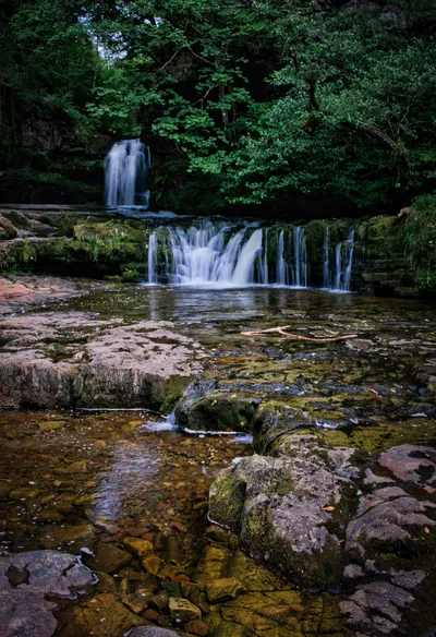 A multi-tiered waterfall cascades over moss-covered rocks in a lush green woodland, with a long exposure effect on the flowing water.