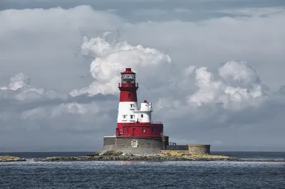 A red and white striped lighthouse on a rocky outcrop in the sea, with a cloudy sky overhead.