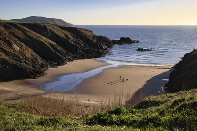 A wide view of Porth Iago beach in Wales, showing two people and a dog walking on the sand, with cliffs on either side and the sea under a golden sky.
