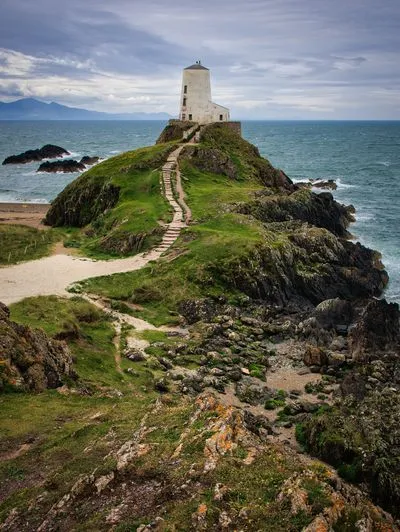 A white lighthouse building stands atop a rocky, green island connected by a winding stone path. The sea with waves crashes against the coast, and mountains are visible in the distance under a cloudy 