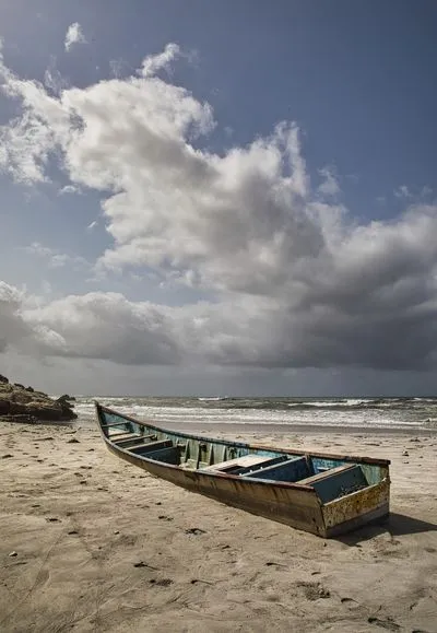 An old, weathered blue boat lies on a sandy beach. In the background, the ocean has white waves, and the sky is filled with dramatic grey clouds. Rocks are visible on the left.