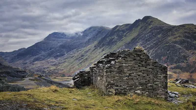 A ruined stone building, likely a slate shed, stands on a grassy hill overlooking a vast mountainous landscape with a quarry and winding roads under a cloudy sky.