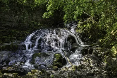 A long exposure photograph of a multi-tiered waterfall flowing over dark, mossy rocks in a lush green forest, with dappled sunlight.