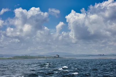 A wide shot from the sea showing the Inner Farne Islands Lighthouse on a small island to the left, and Bamburgh Castle on the mainland to the right, under a cloudy sky.