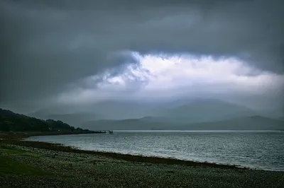 A landscape view of a pier extending into a body of water, with a rocky shore in the foreground, forested hills on the left, and misty mountains under a dark, cloudy sky.