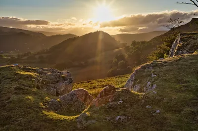 A landscape photograph of Dinas Bran castle silhouetted against a bright sunset with sunbeams over a valley of green hills and grassy rocks in the foreground.
